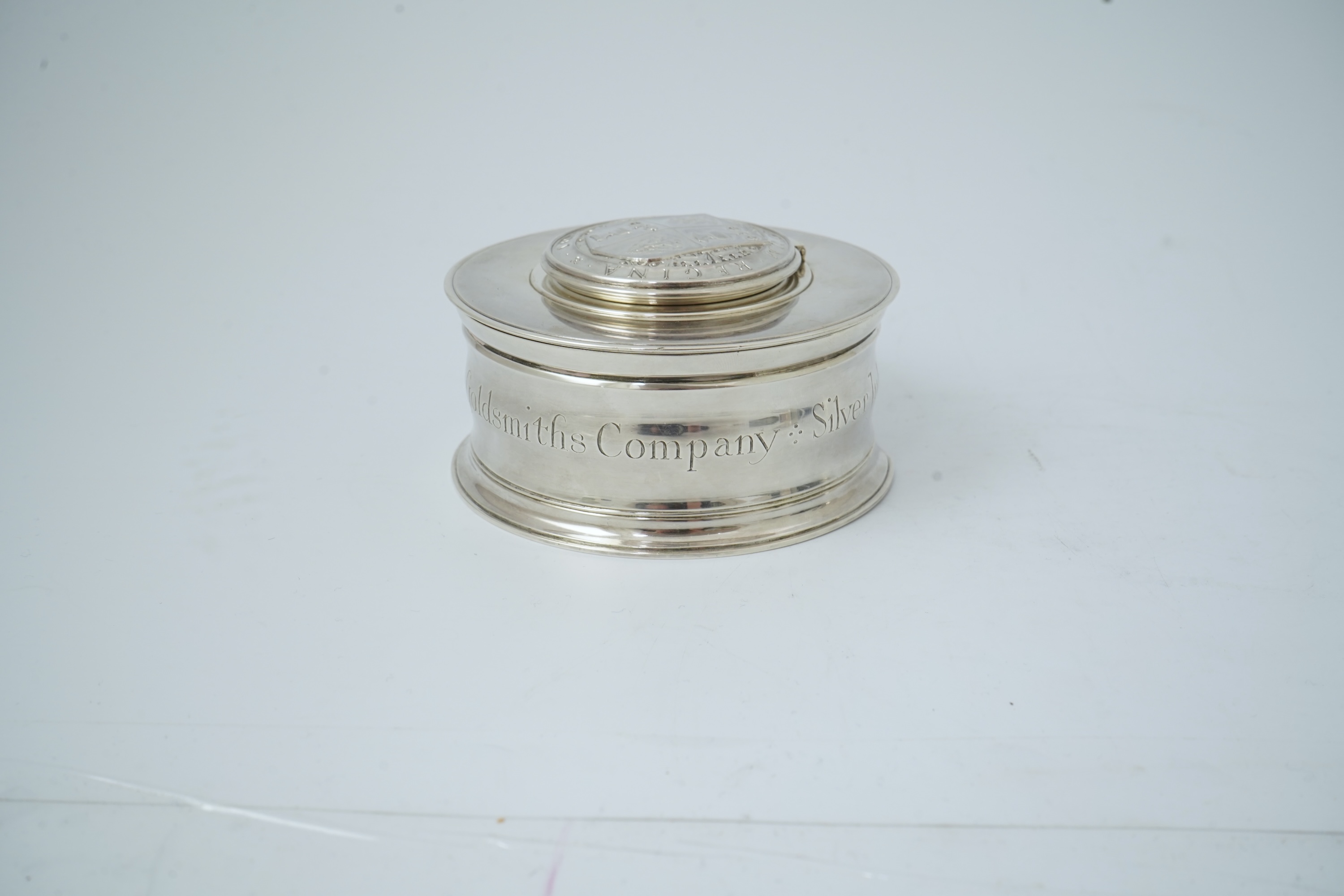 A George V silver circular inkwell, by Sebastian Garrard, commemorating the Silver Jubilee of King George V, the die stamped cover designed by George Kruger Gray featuring the Goldsmiths Company Coat of Arms